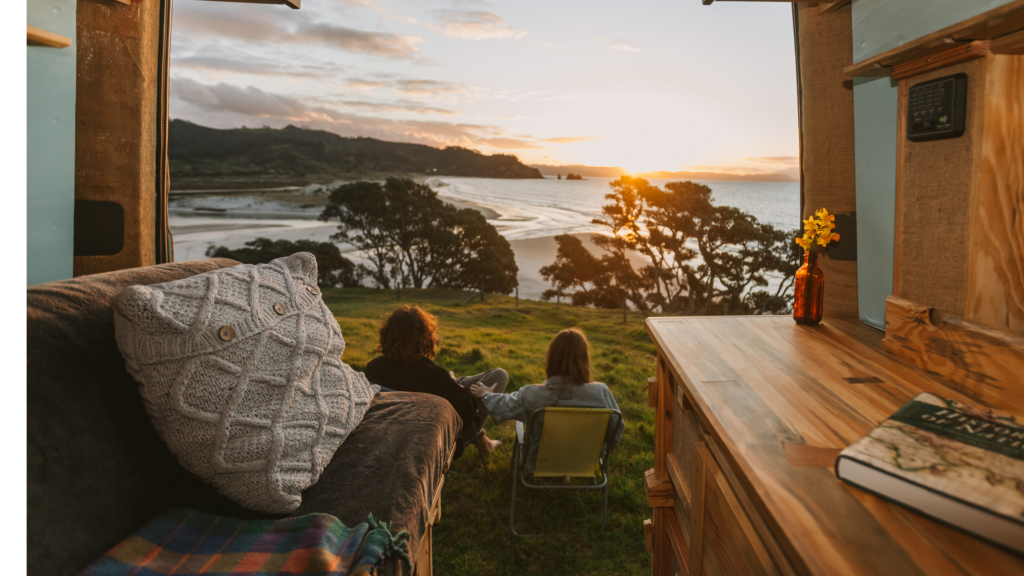 A couple sit together outside their camper taking in the sunset by the beach on their campervan holiday in new zealand
