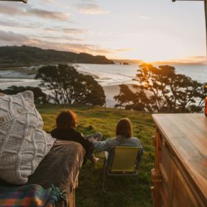 Looking out rear of campervan, a couple sit taking in a view of sunset over beach and sea