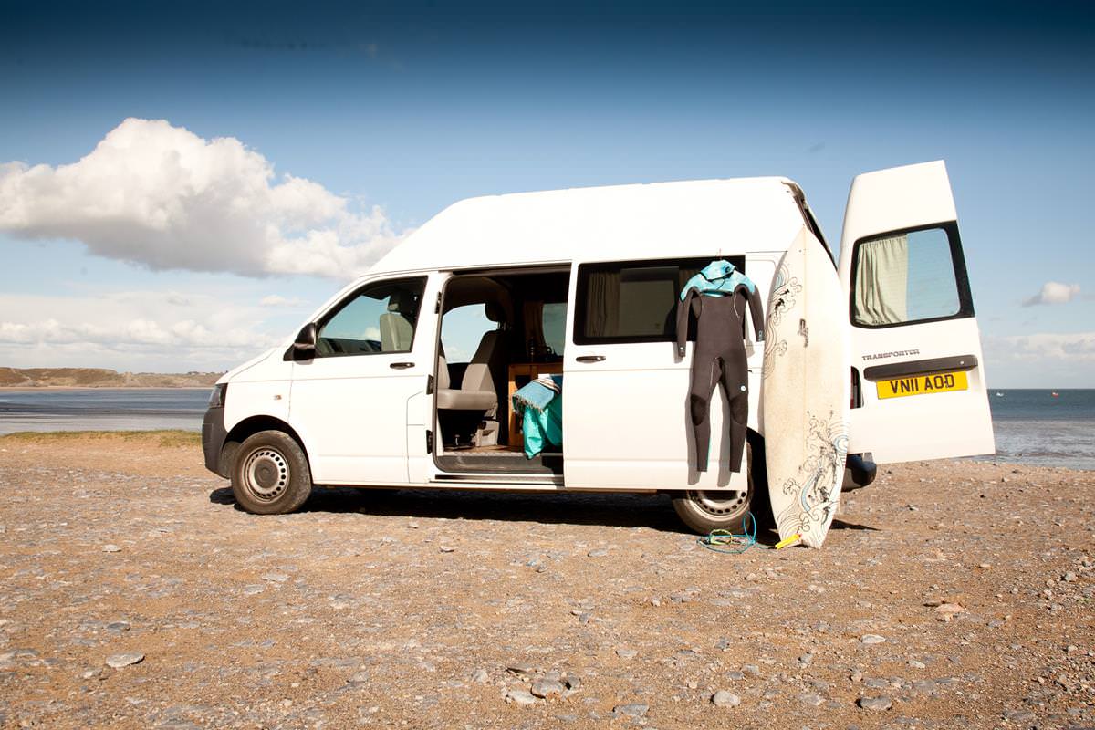 Sandy the campervan on the beach with a surfboard and wetsuit