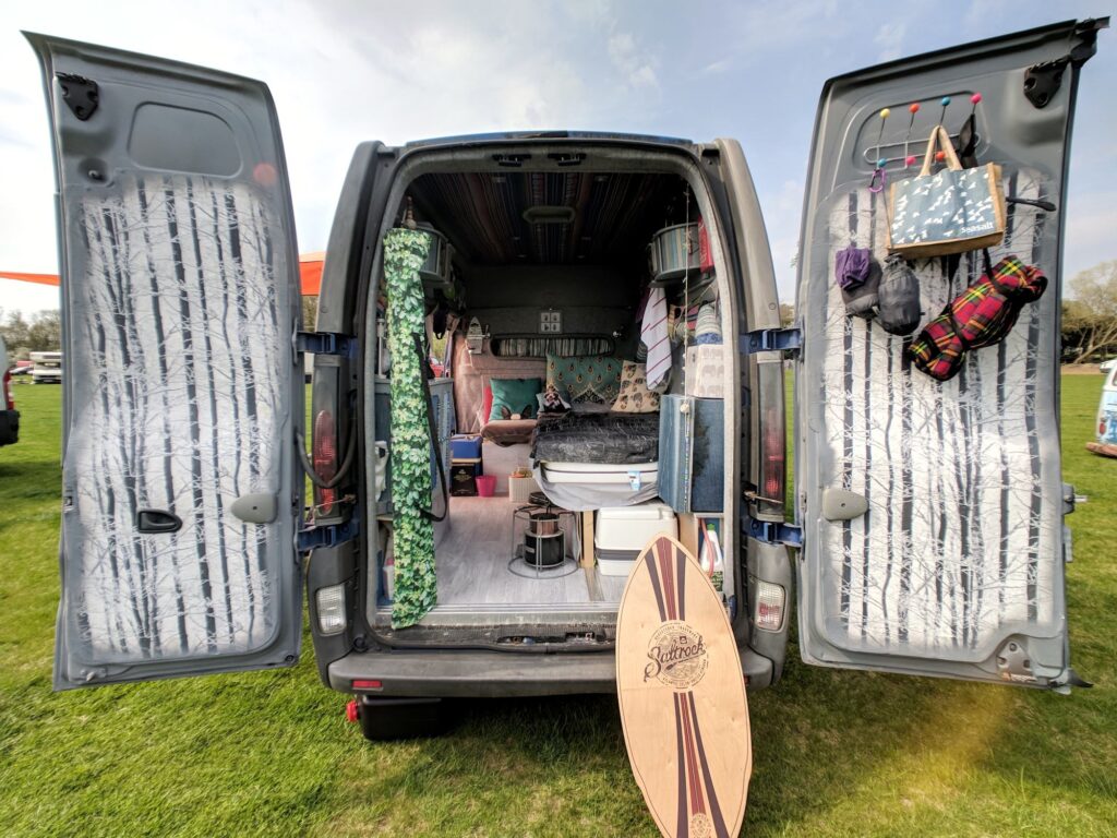 The back doors open of a selfbuilt campervan showing the living area, picnic banket and skateboard