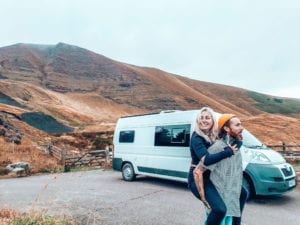 A man giving a lady a piggyback in front of their campervan in the mountains