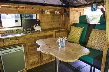 A cozy van interior with wooden cabinetry and a small fridge. A live-edge wooden table with a vase of white and purple flowers is set in front of a cushioned seating area. The kitchen area includes a stove, a kettle, and hanging mugs, accented by a window showing greenery outside.