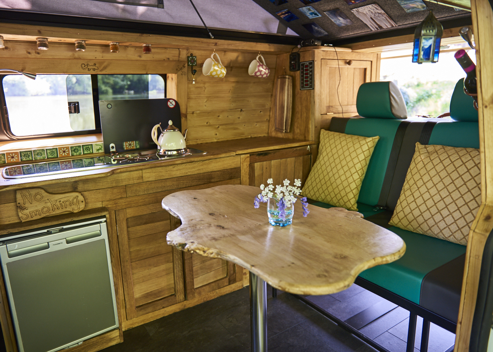 A cozy van interior with wooden cabinetry and a small fridge. A live-edge wooden table with a vase of white and purple flowers is set in front of a cushioned seating area. The kitchen area includes a stove, a kettle, and hanging mugs, accented by a window showing greenery outside.
