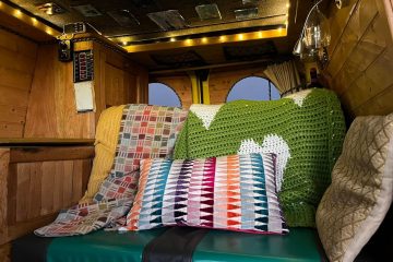 The image shows the cozy interior of a camper van. The seating area features a blue-green cushioned bench, draped with a green crochet blanket and a yellow quilt. A colorful geometric-patterned pillow rests on the bench. The wooden walls and soft lighting create a warm, inviting atmosphere.