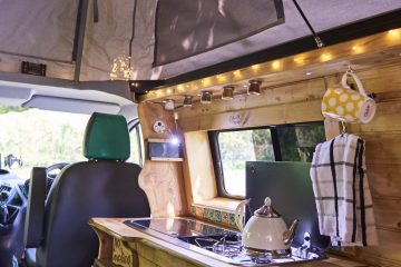 Interior of a camper van showcasing a compact kitchen. It features a stovetop with a kettle, a wooden countertop, spice jars mounted above, a checkered towel hanging, and a yellow polka-dotted mug. The van has green seat covers and is well-lit with both natural light from windows and string lights.