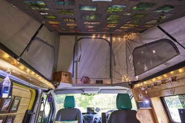 Interior of a camper van with cozy decorations. The ceiling is adorned with photographs. The walls are decorated with wooden accents and small jars. Fairy lights illuminate the space, providing a warm ambiance. The seating area includes green and black seats with pillows.