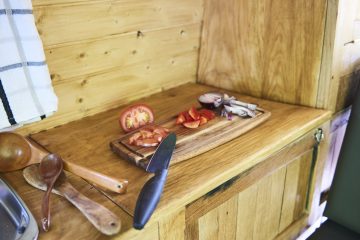 A wooden kitchen counter with a cutting board holding chopped tomatoes and onions. Several wooden utensils and a black-handled knife rest beside the chopping board. A white and black checkered towel hangs on the wall above the counter, and a wooden cabinet door is visible below the counter.