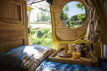 A cozy yellow campervan interior with wooden finishes. There's a bed draped with a blue blanket, and a tray on it that holds a French press, a glass of juice, and a polka-dotted mug. Through the large circular window, a sunny, green, grassy landscape with trees is visible.