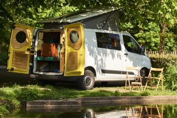 A white camper van with open yellow rear doors is parked by a lakeside, partially shaded by trees. Two wooden chairs are set up outside the van, facing the water. Inside, the van features a cushioned seating area and light wooden interior elements. It's a sunny, serene outdoor setting.