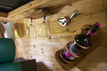 A close-up of a wooden interior wall of a camper van showcasing a compact wine storage solution. Two wine glasses hang upside down from a wooden rack, and a wine bottle is secured in a wall-mounted wooden holder. The background features light-colored wooden paneling and a metal hook.