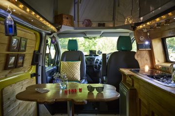 Interior of a cozy, well-lit camper van. The wooden table holds sunglasses, a candle, red dice, and a small vase with flowers. The front seats are upholstered in gray and teal. The cabinets and walls are wooden, adorned with string lights, small pictures, and miscellaneous items. Trees are visible through the front windshield.
