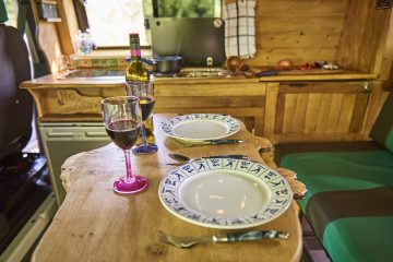 A cozy dining setup inside a camper van with a wooden table set for two. The table features two plates, utensils, and two glasses of red wine. The kitchen area in the background includes a counter with cooking utensils, a bottle of wine, and a "No Smoking" sign. The seating area has green cushions.