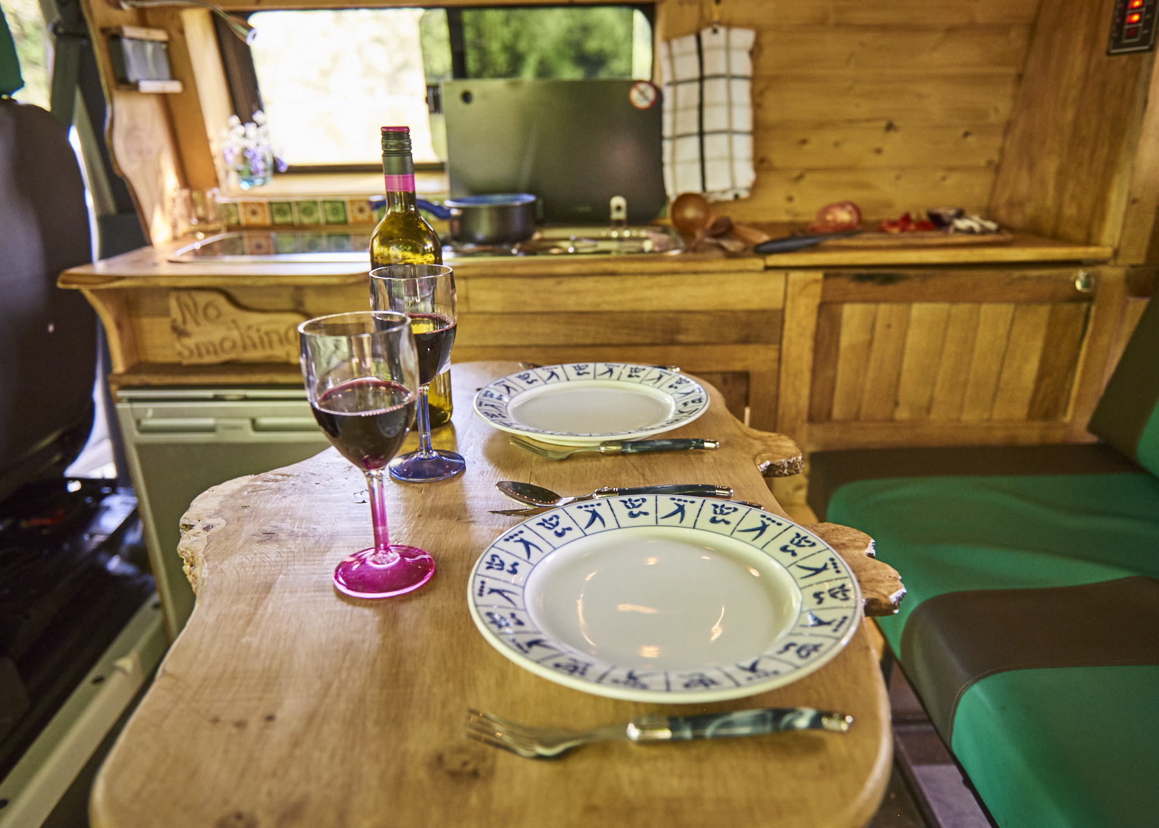 A cozy dining setup inside a camper van with a wooden table set for two. The table features two plates, utensils, and two glasses of red wine. The kitchen area in the background includes a counter with cooking utensils, a bottle of wine, and a