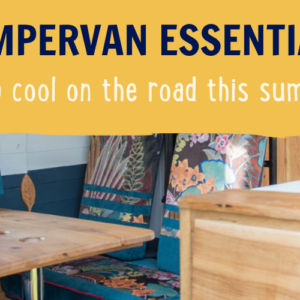 Interior of a campervan with a wooden table and benches covered in colorful cushions with floral patterns. Above the seating area, there's a wooden cabinet. The text overlay reads "CAMPERVAN ESSENTIALS keep cool on the road this summer" in bold letters.