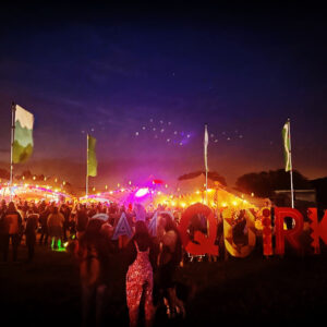 A vibrant outdoor festival scene at dusk, featuring a large crowd gathered near tents adorned with string lights. The foreground features large, colorful letters spelling "CAMP QUIRK." Tall flags on poles surround the area. The sky transitions from deep blue to a warm hue on the horizon.