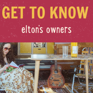 A woman in sunglasses and a floral dress sits reading a book at a wooden table outside a camper van. The table holds a bottle of wine, a glass, and a notebook. The text "GET TO KNOW elton's owners" is overlaid in bold yellow and white letters against a red background at the top.