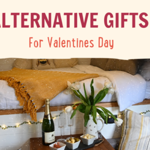 A cozy setting featuring a comfortable bed with white and yellow bedding and a mustard throw. A wooden table holds a bouquet of white tulips, a bottle of sparkling wine, two glasses, and various snacks. Text reads "ALTERNATIVE GIFTS For Valentines Day" in warm tones. String lights add a soft glow.