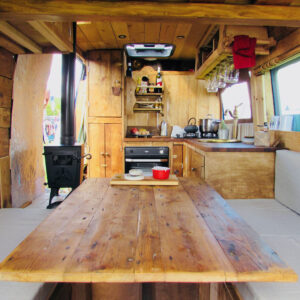 The interior of a rustic wooden camper van includes a central wooden table with benches on either side, a kitchen area with a stove, sink, and shelves filled with kitchenware. A small wood-burning stove is on the left, and a window on the right allows natural light to illuminate the space.