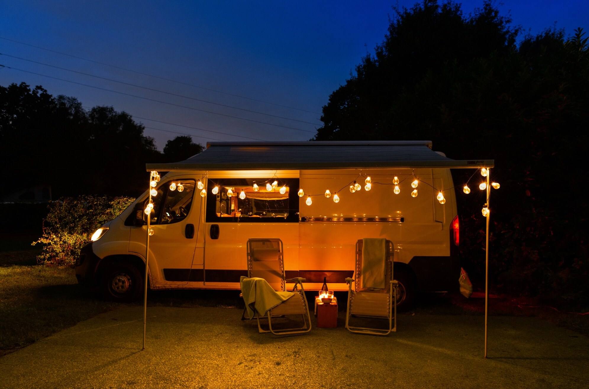 A campervan with string lights parked at dusk, with two chairs and a small glowing table beneath its awning.