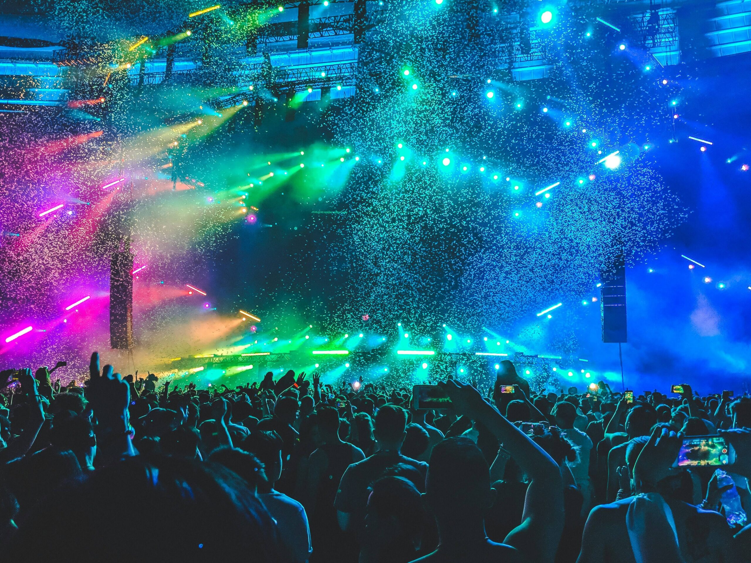 A crowd raises hands and phones toward a concert stage filled with blue, green, pink, and yellow lights and falling confetti.