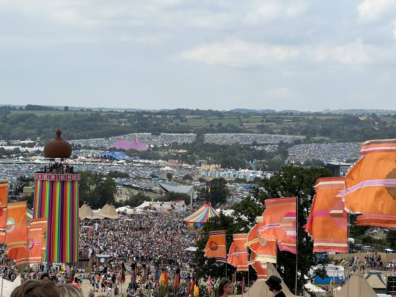 A crowded festival fills a hillside, with colorful striped flags, tents, and fields of parked cars stretching across the landscape