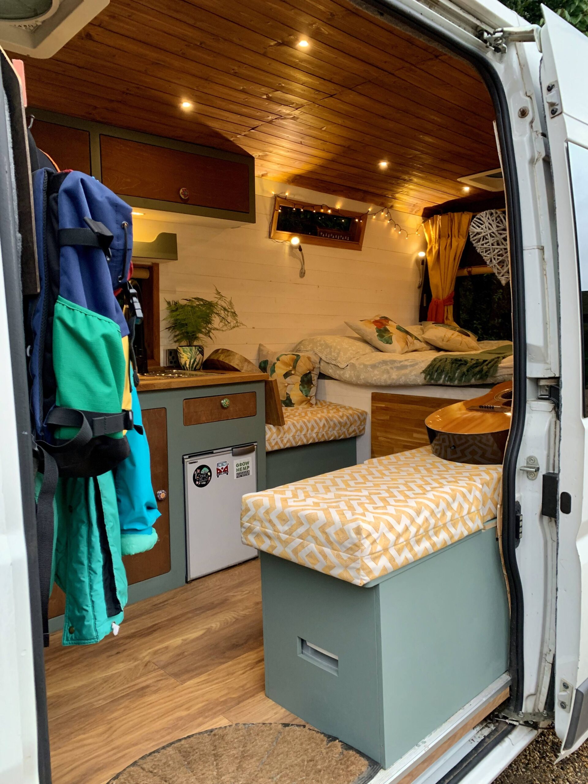 View into a campervan through the side door with warm lighting. Showing the bed, a portion of the kichen and the seating area