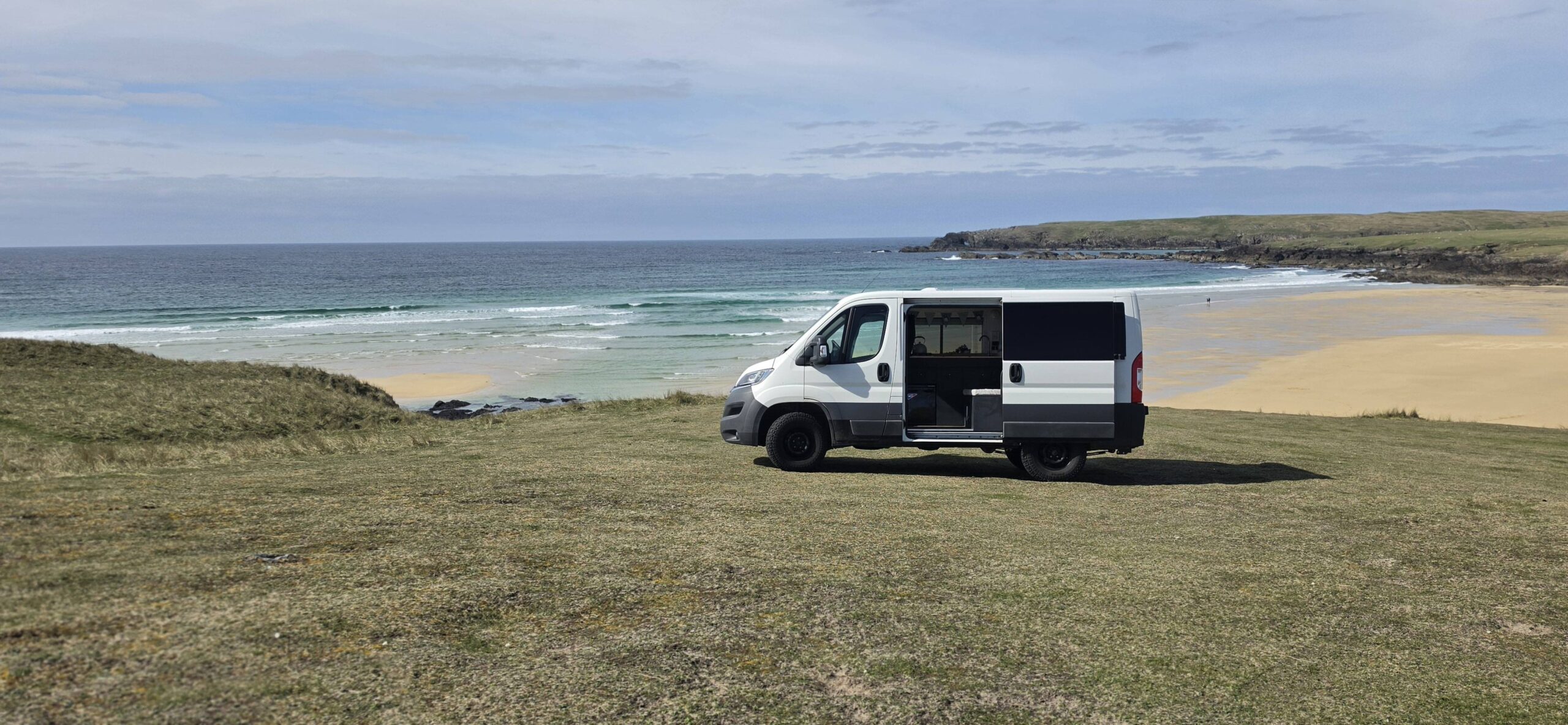 Compact campervan parked on a grass verge with a beach and ocean in the back ground