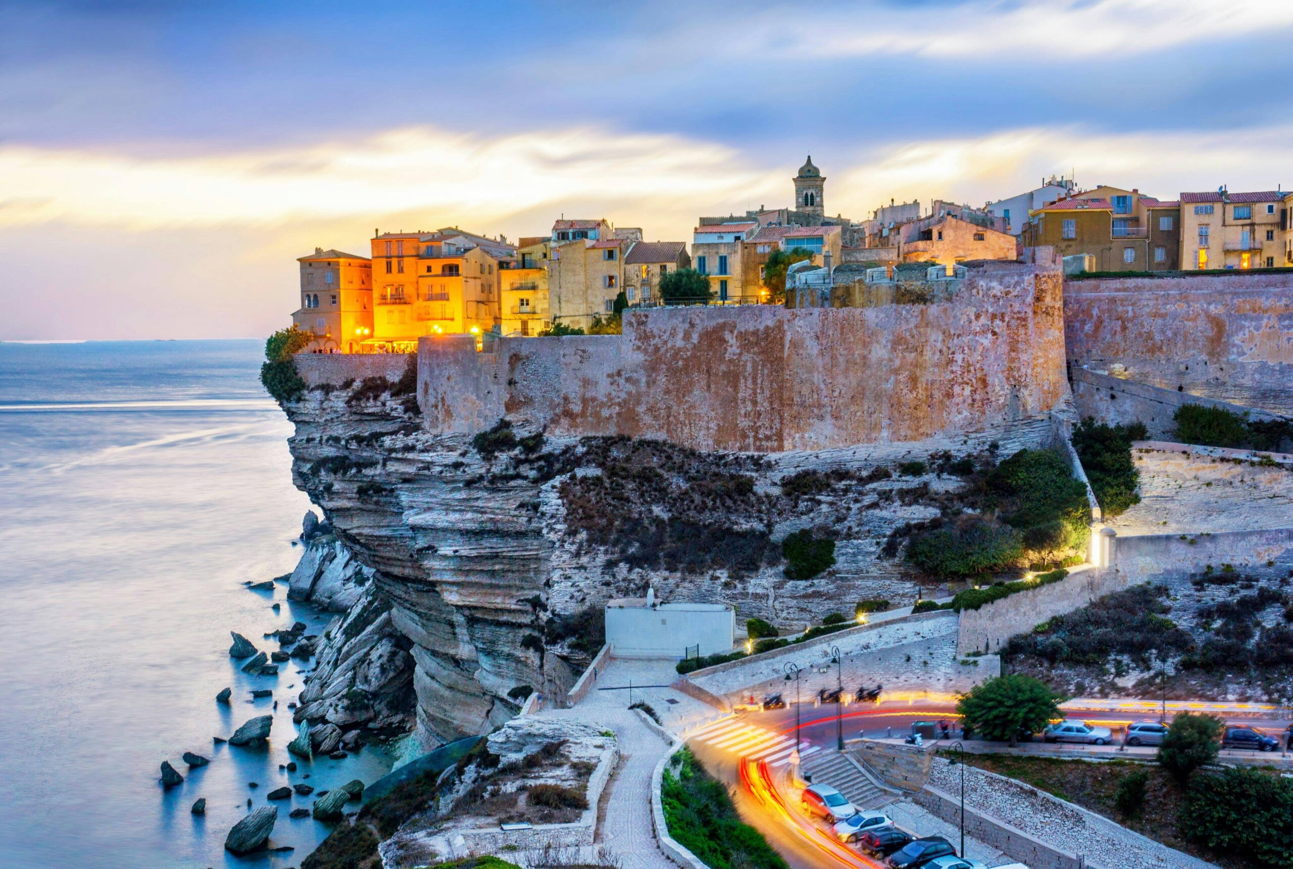 Colorful buildings sit atop high coastal cliffs overlooking the sea at dusk, with a winding road and streaks of car lights visible below.