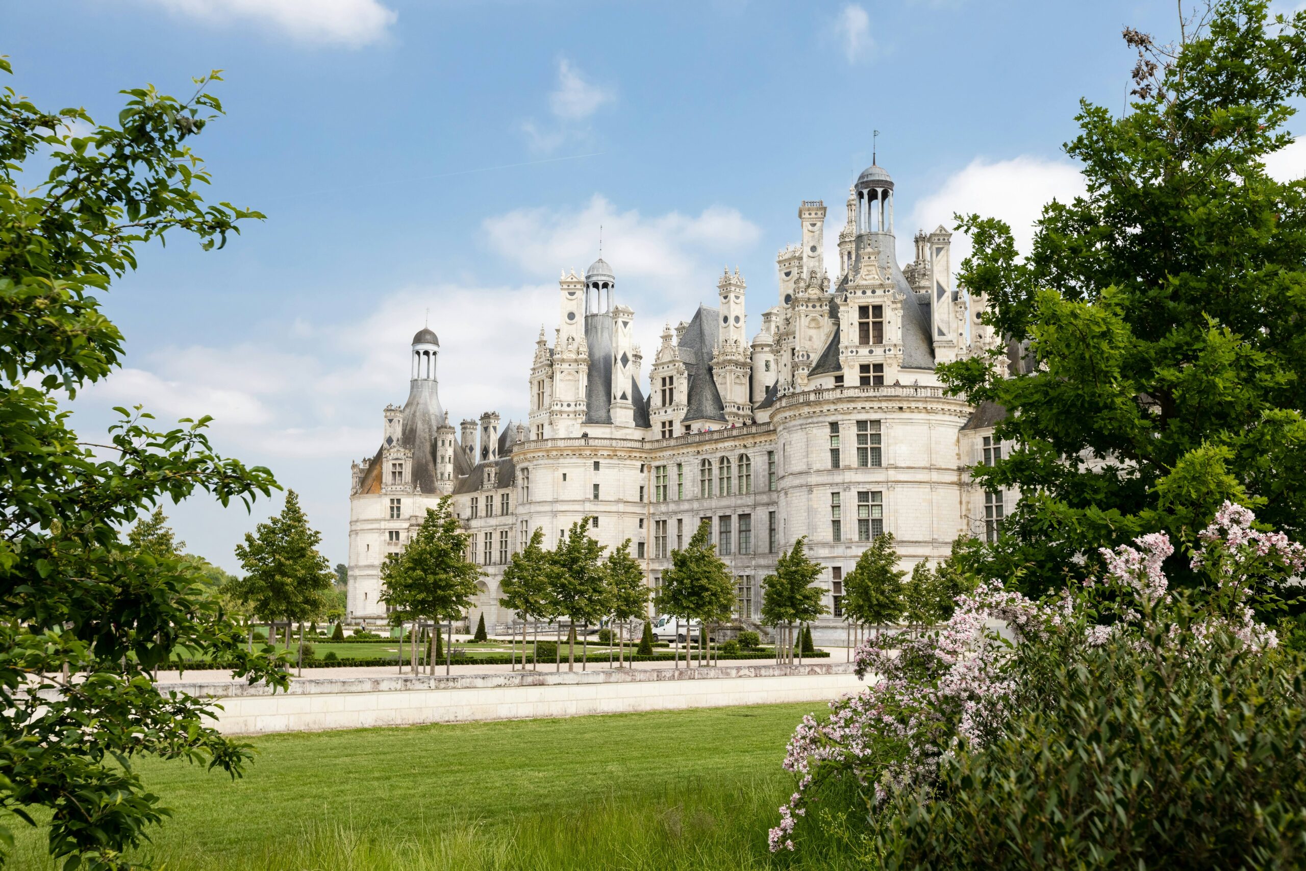 A grand historic château surrounded by lush greenery and trees, with intricate architectural details and a clear blue sky in the background.