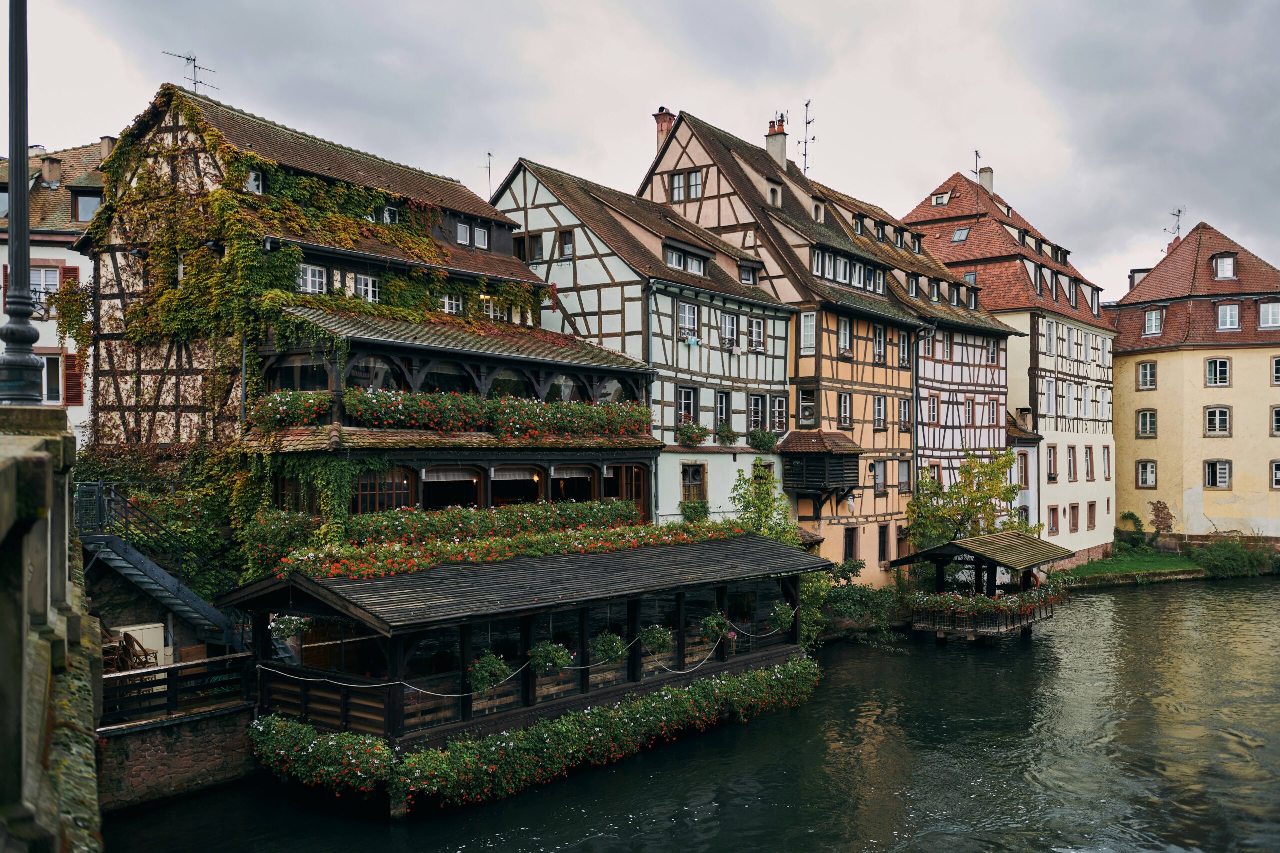A row of historic timber-framed buildings lines a canal, with lush greenery and outdoor seating areas overhanging the water under a cloudy sky.