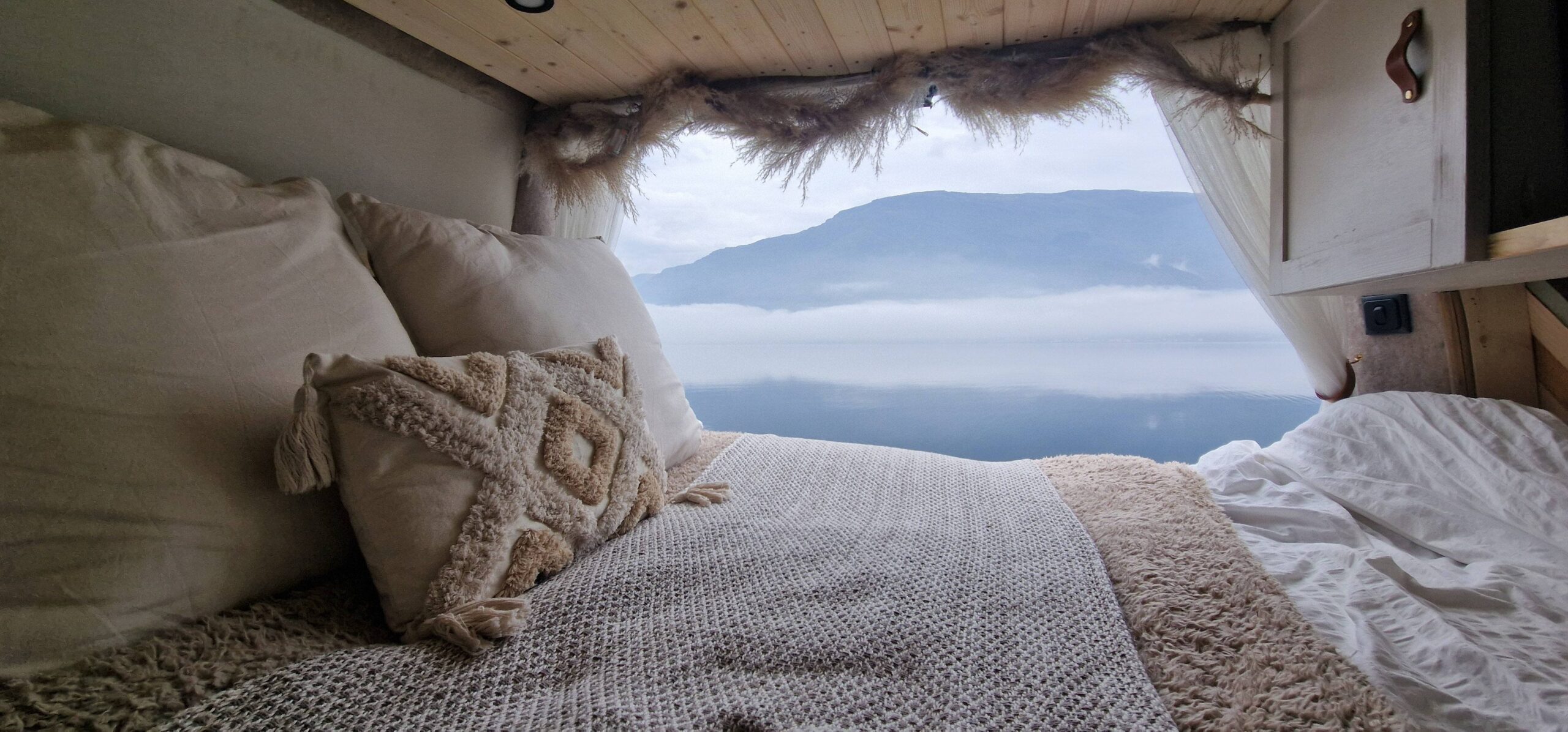 A cozy bed with soft pillows and a textured blanket, situated in a space with the rear doors open, revealing a misty mountain landscape outside.
