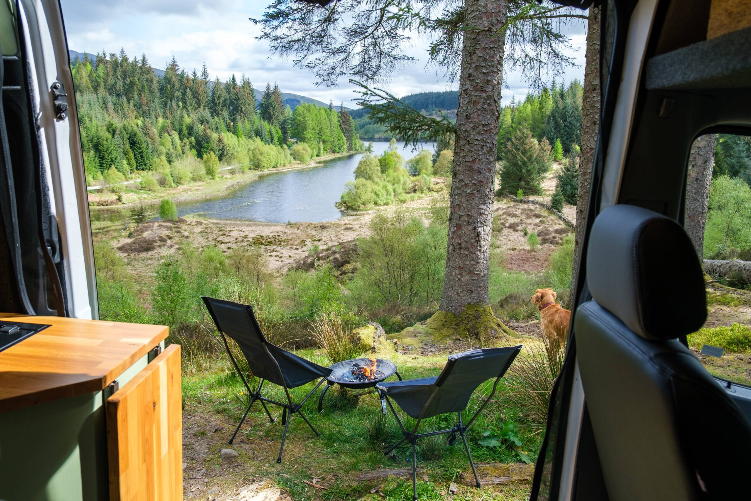 A scenic view from inside a van, showing a river surrounded by trees and greenery, with two chairs and a small table set up outside.
