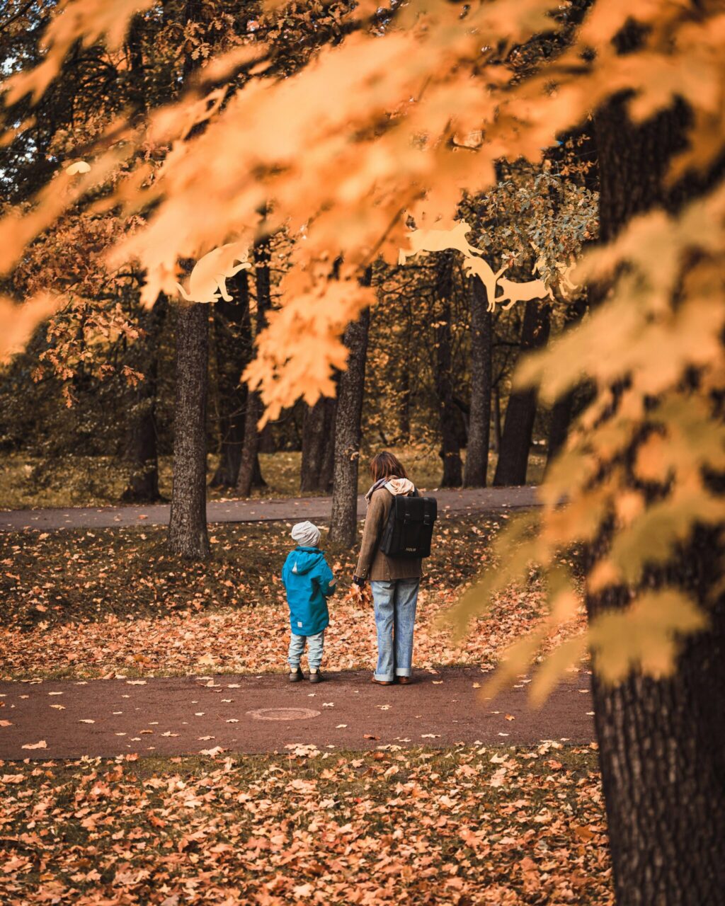 A parent with a backpack walks hand in hand with a child along a leaf-strewn autumn park path, framed by golden-orange foliage; the mood is peaceful.