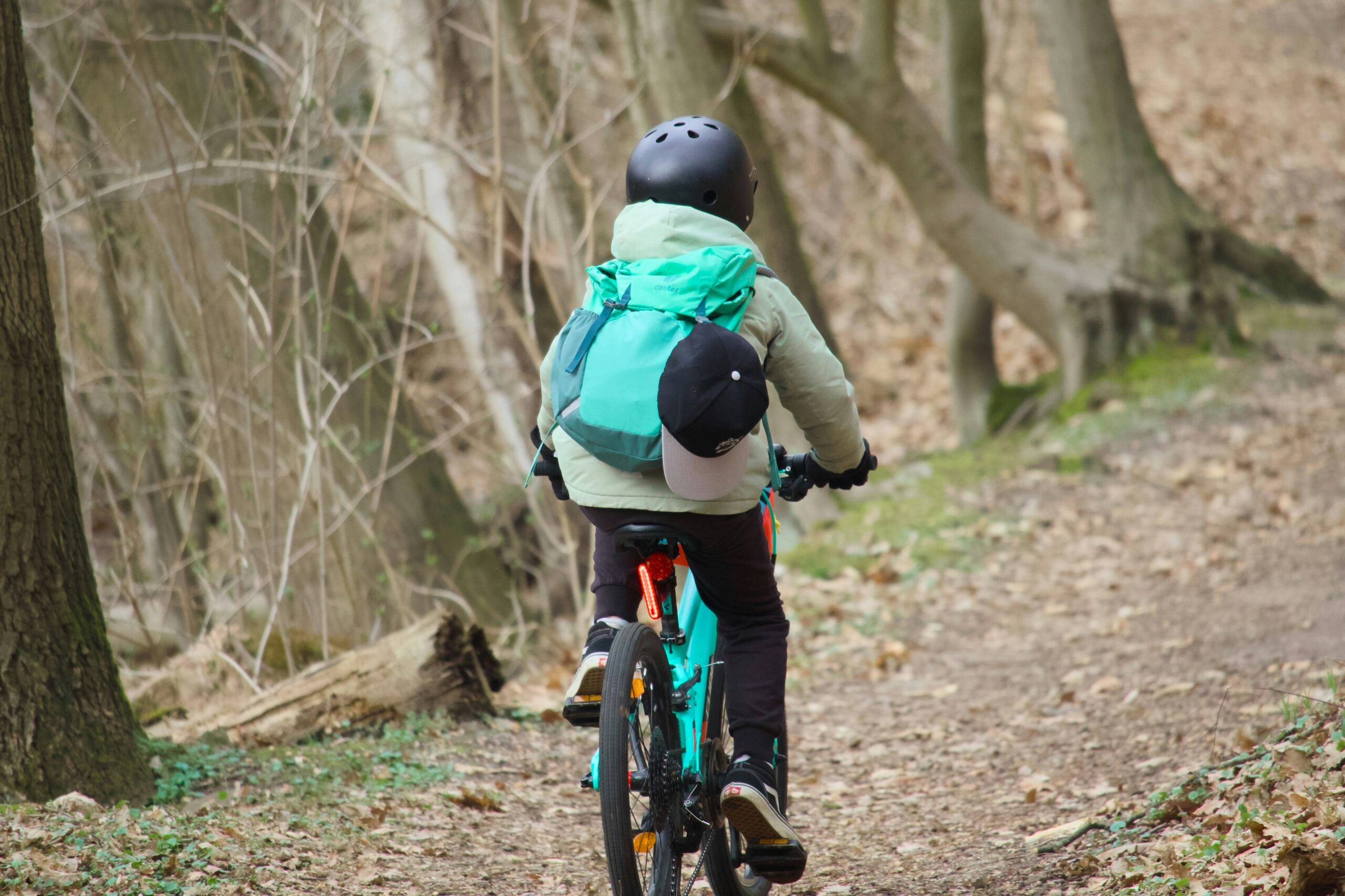 A helmeted cyclist with a bright teal backpack rides a turquoise bike along a quiet, leaf-strewn woodland trail, conveying calm adventure.