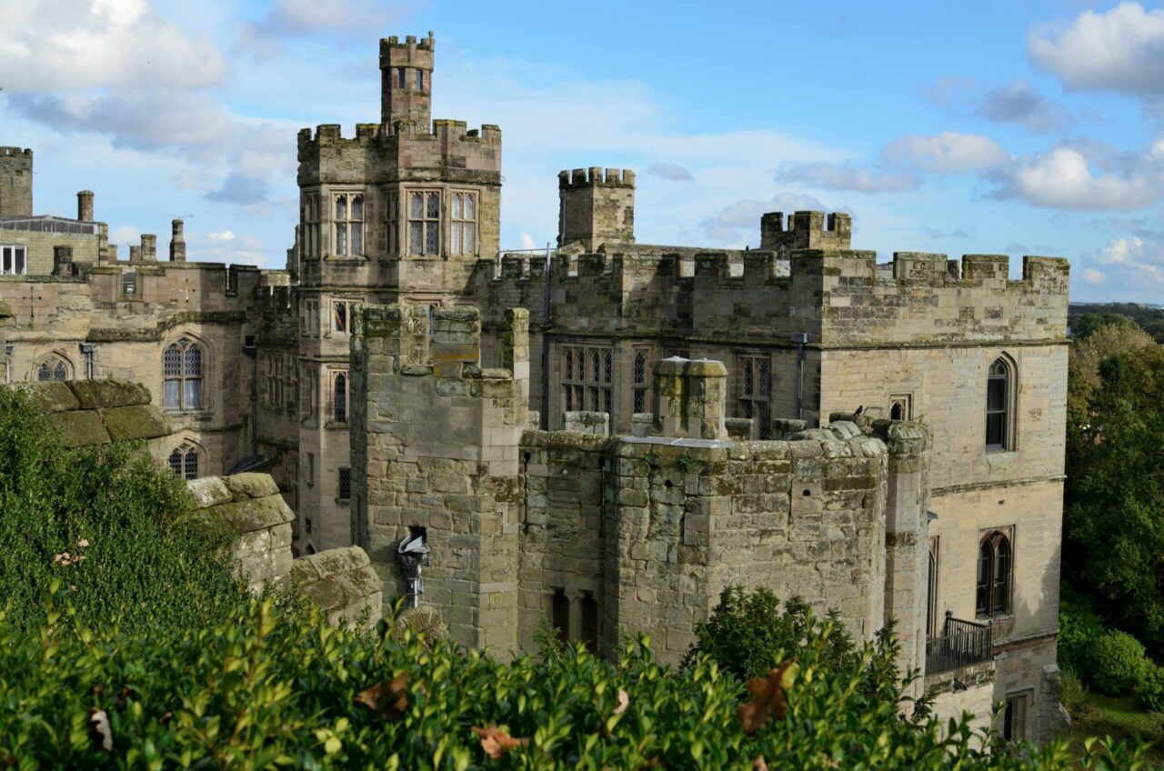 Historic stone castle with crenellated towers and arched windows rises above green shrubs beneath a bright, cloud-dappled sky, conveying a timeless, peaceful grandeur.