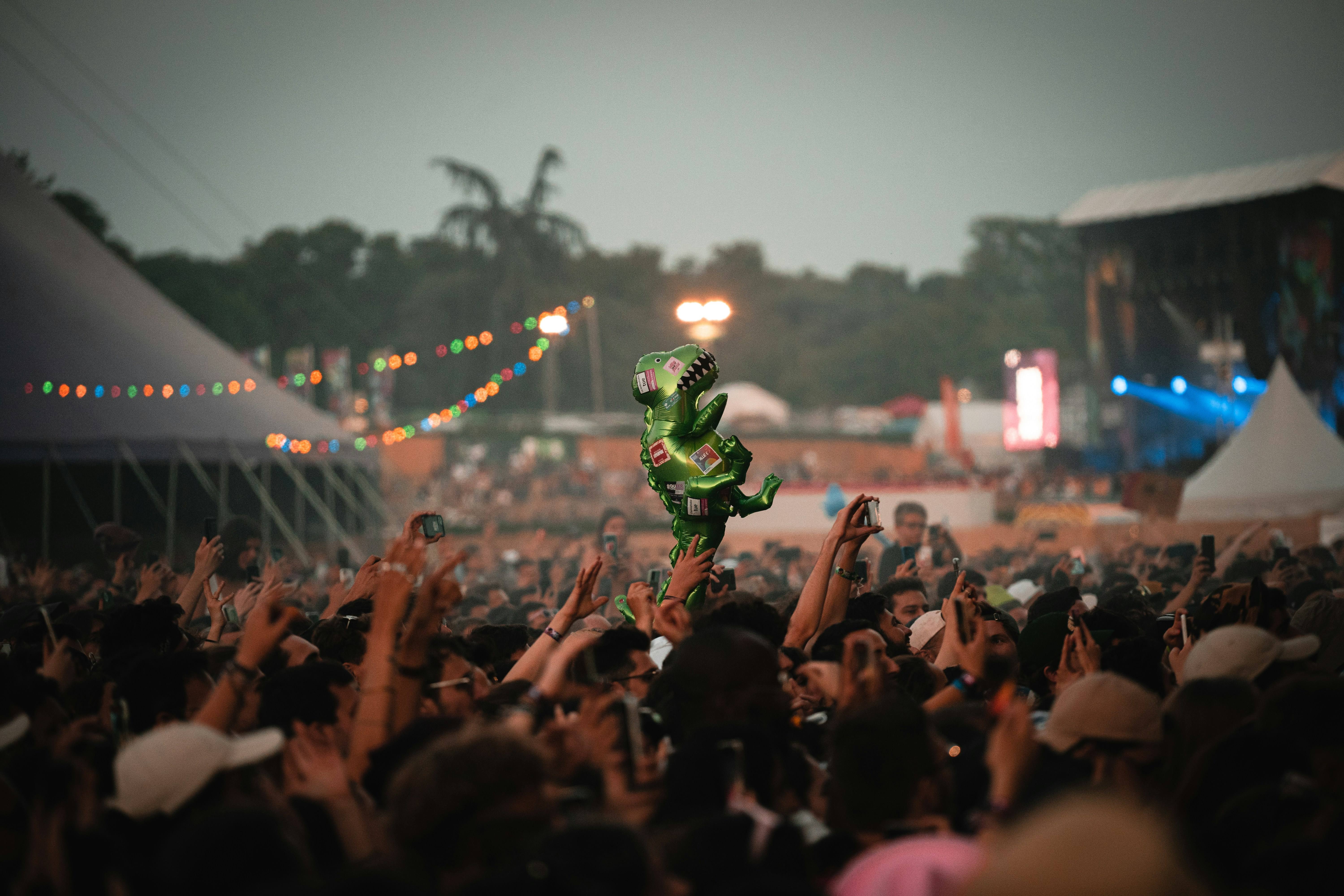 A lively festival crowd raises hands as a green inflatable dinosaur floats overhead, with colorful string lights and tents glowing at dusk.