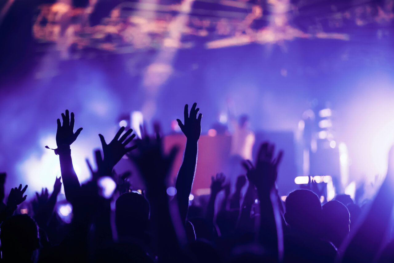 Silhouetted concert crowd raises hands beneath purple and blue stage lights, creating an energetic, euphoric atmosphere at a live show.