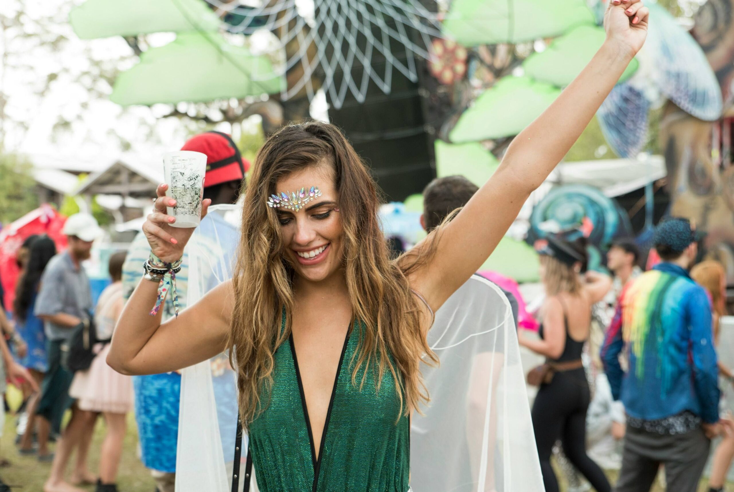 Smiling woman in glittering green outfit dances at a lively outdoor festival, raising one arm and holding a drink amid colourful crowds.