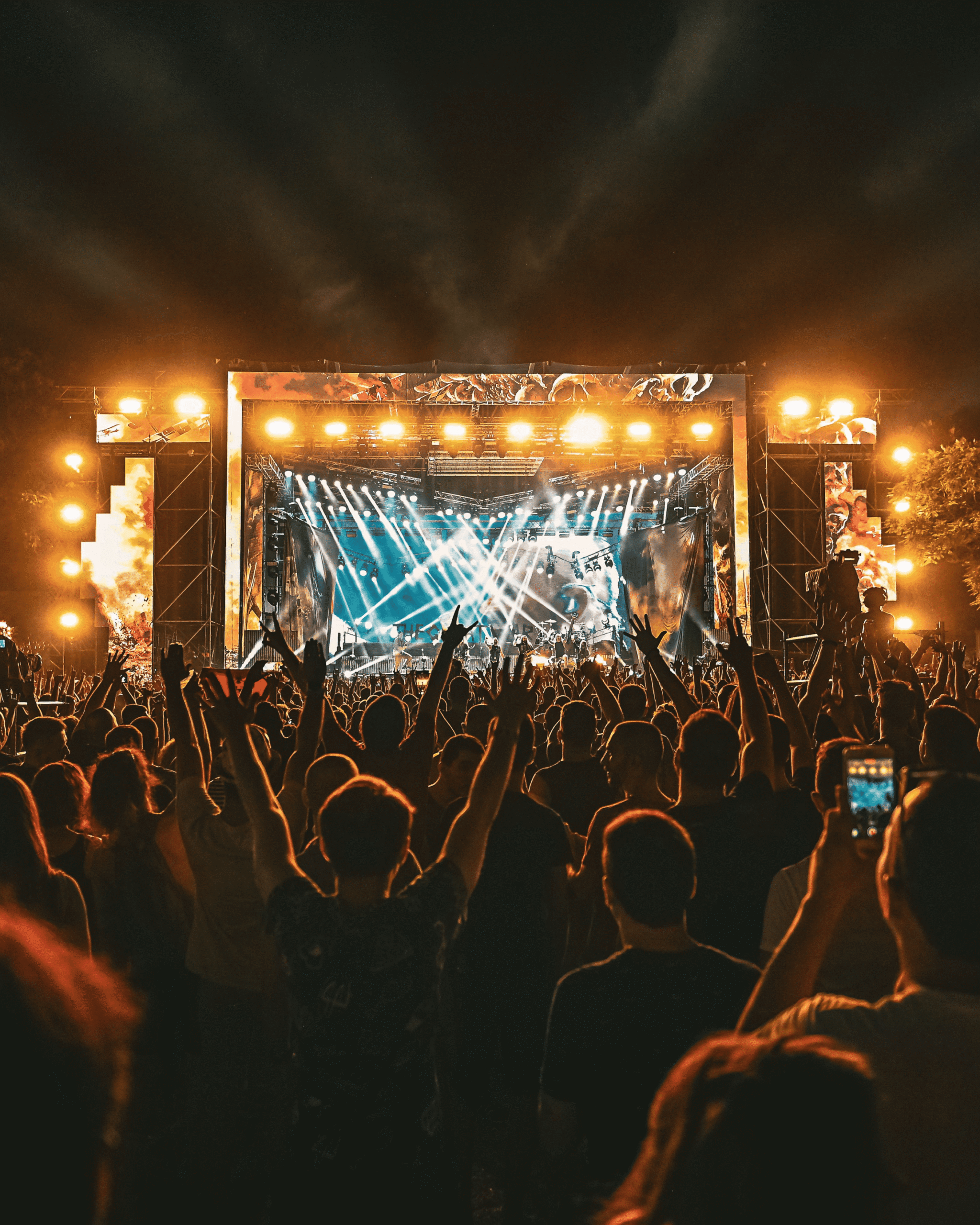 A large crowd faces a brightly lit outdoor concert stage at night, with people raising their hands and taking photos as beams of light shine across the audience.