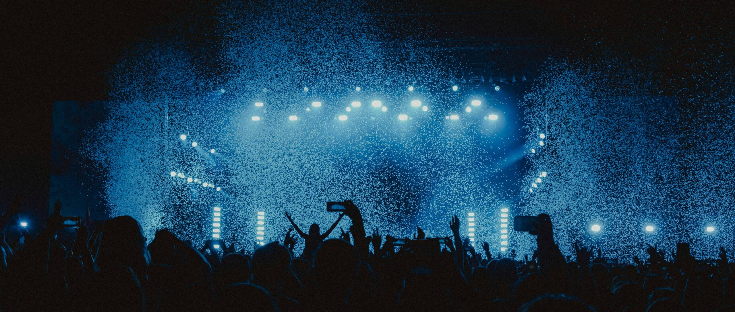 Concert crowd silhouetted with raised hands beneath bright blue stage lights and mist, creating an energetic, immersive festival atmosphere.