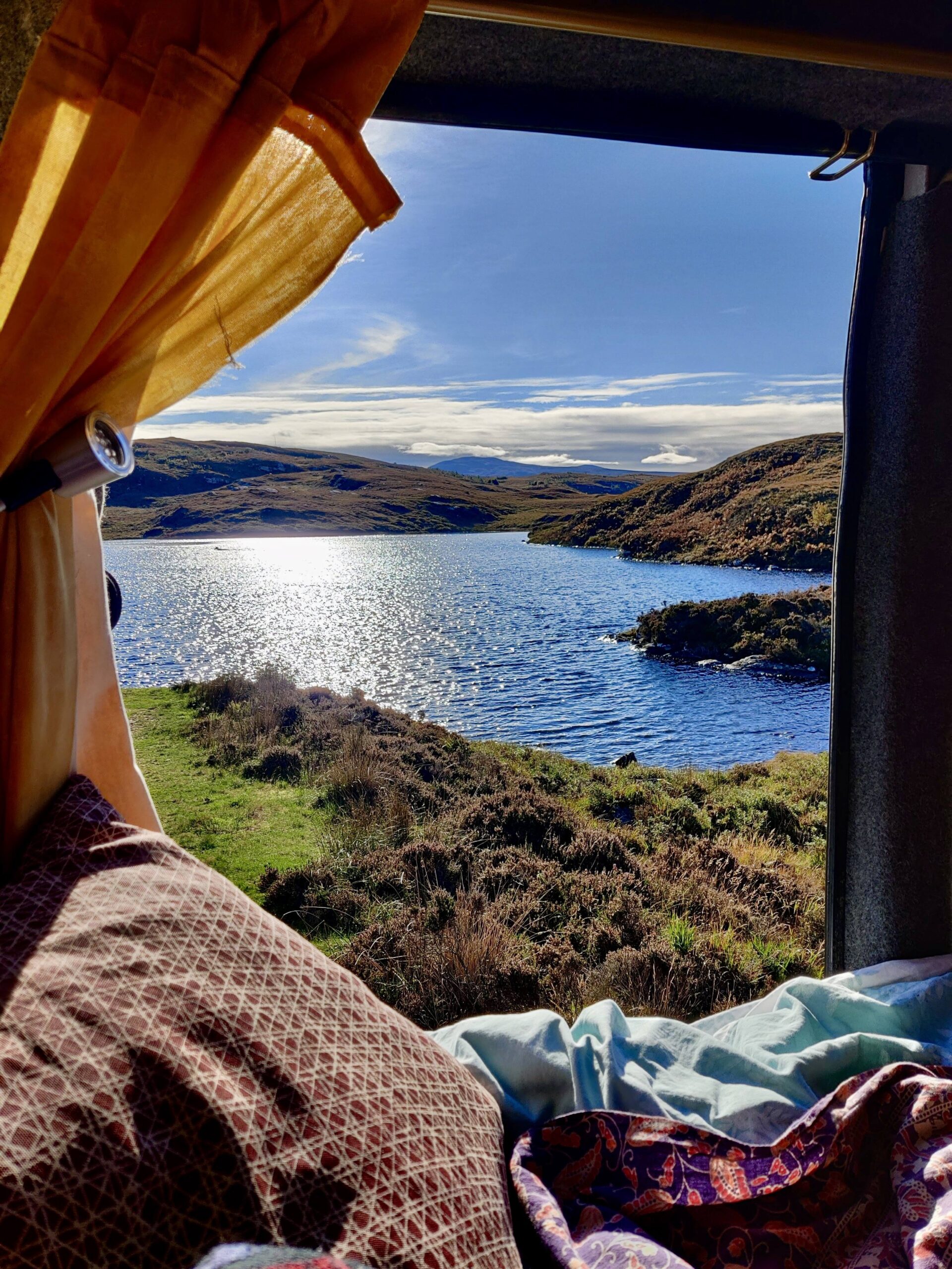 The view from the open back door of a camper. Showing a lake glistening in the sun with clear blue skies.