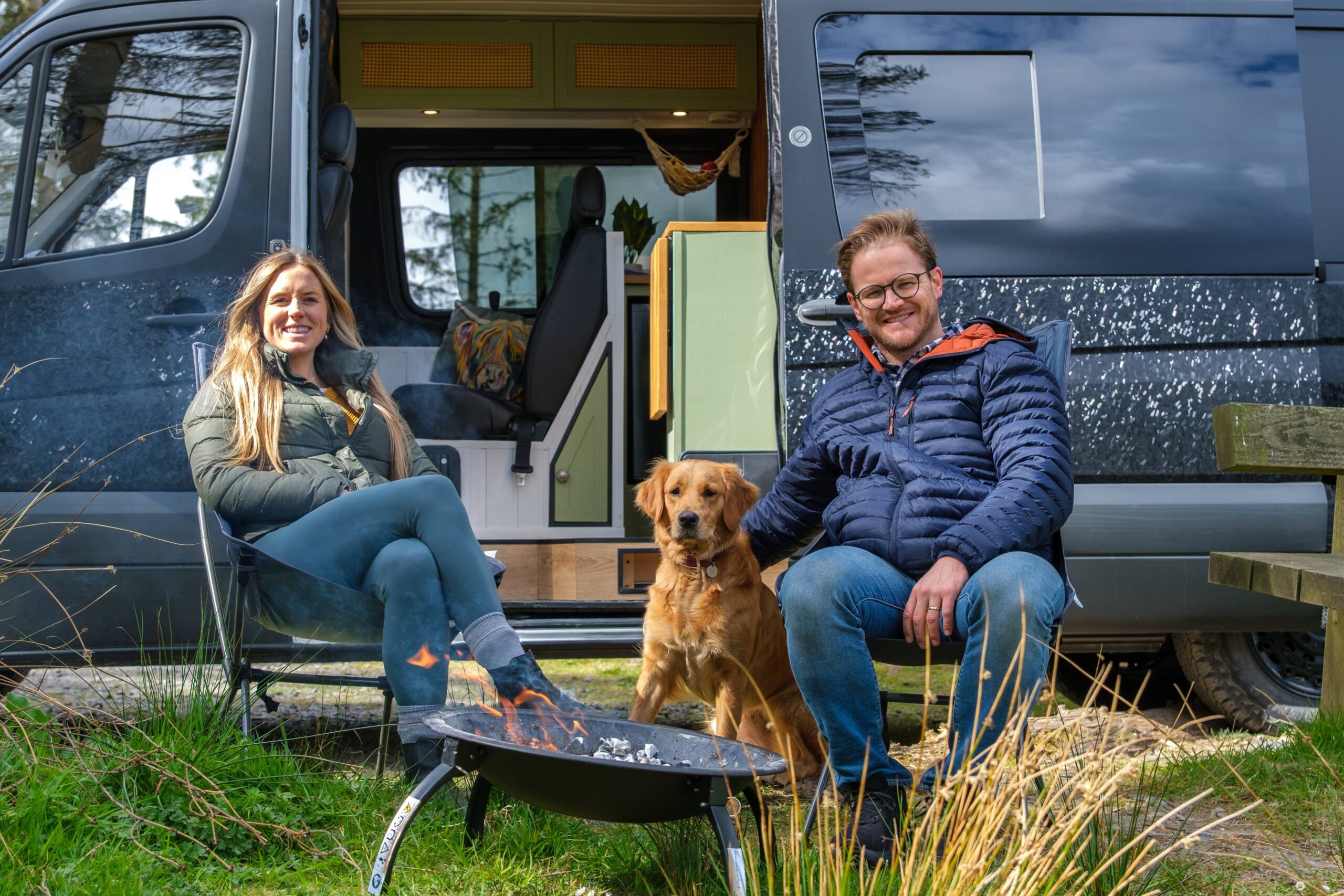 Two happy campervan owners sat out side of the campervan. Between them sits a dog.