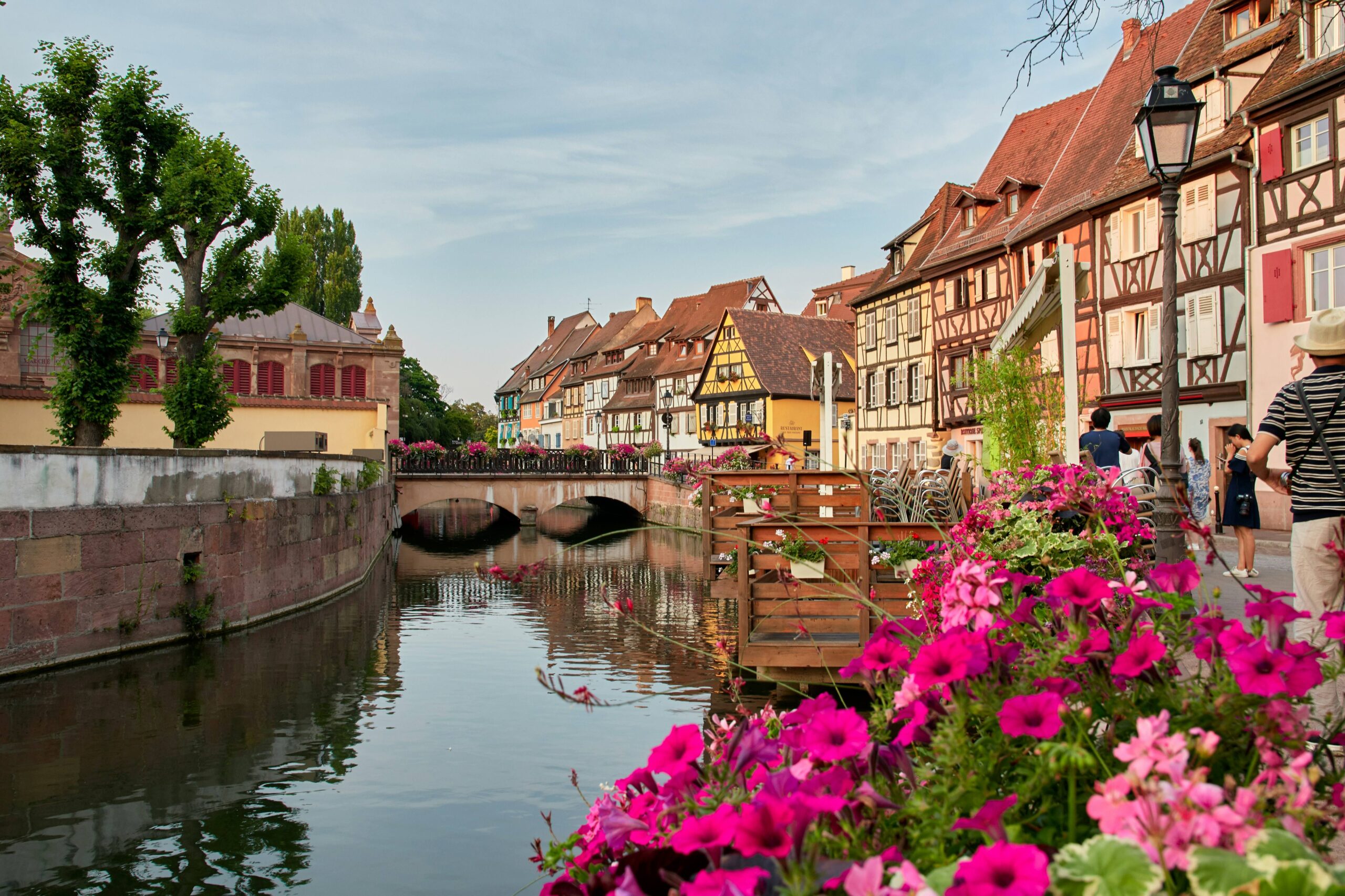 A canal flows through a historic town with traditional half-timbered houses, a stone bridge, and vibrant pink flowers blooming along the waterfront.