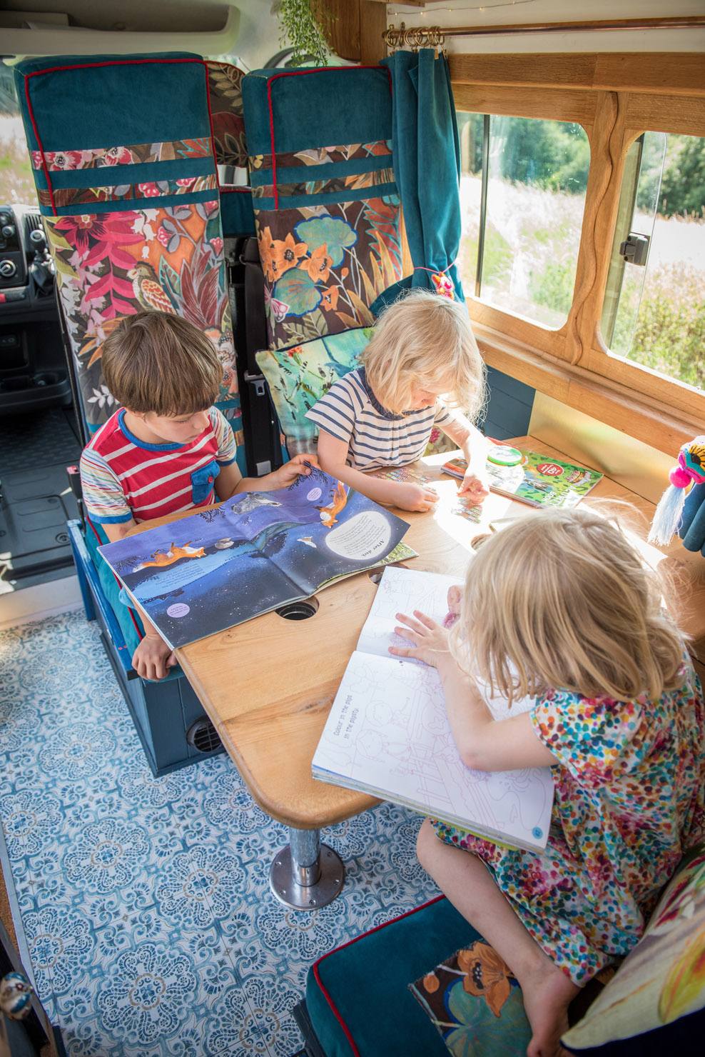 Three children sat at the table inside of a campervan. The children are reading books and colouring.