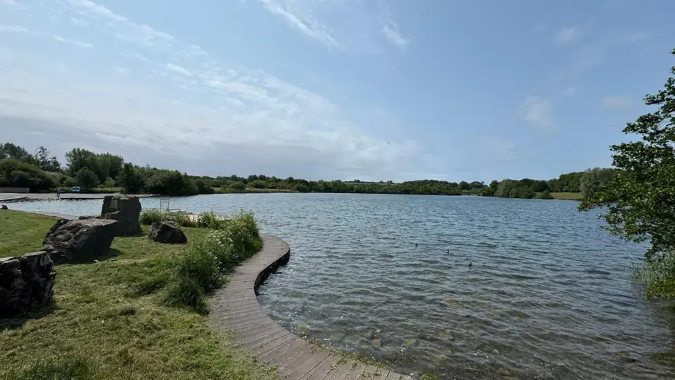 A serene lake scene featuring a wooden walkway leading to a grassy area, surrounded by trees and calm water.