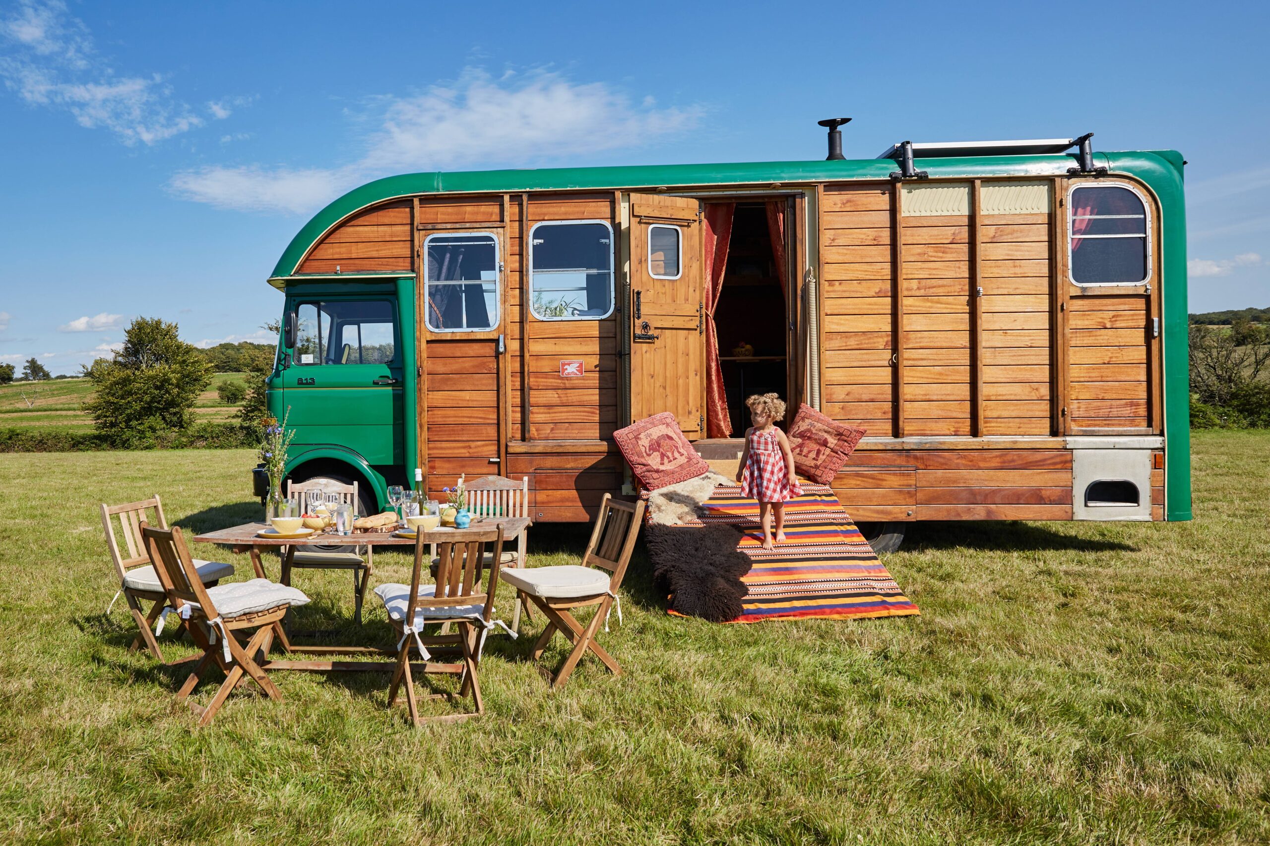 A large horse box campervan with the side door open. Outside is a small child with a picnic table to their left