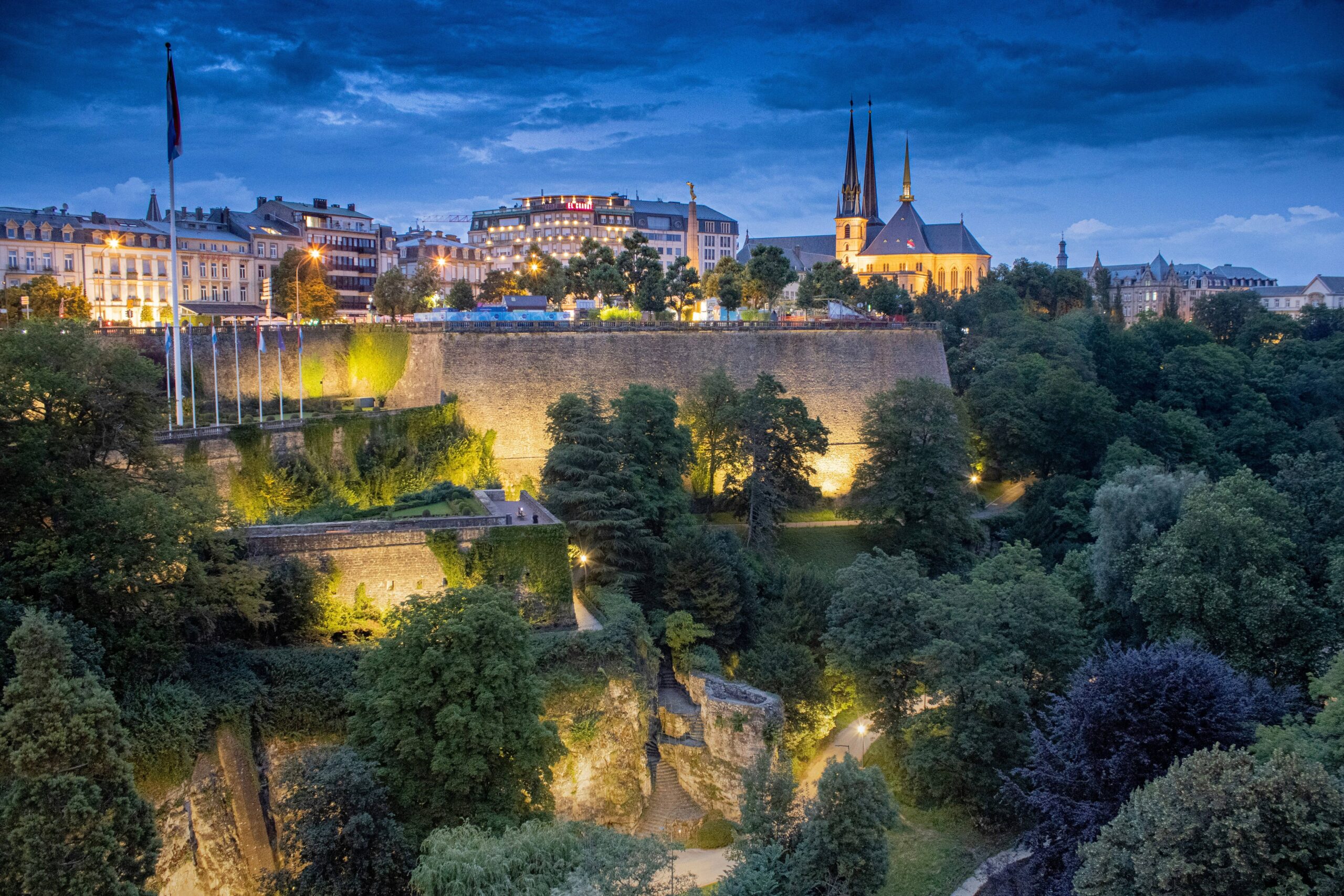 Panoramic view of Luxembourg City at twilight, featuring historic stone fortifications, lush green trees, and the spires of the Notre-Dame Cathedral.