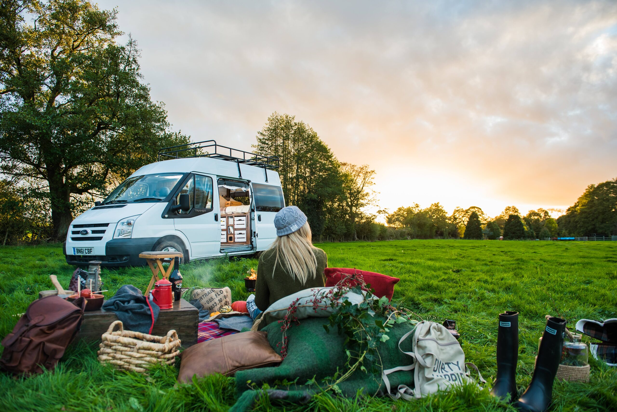 A campevan parked up with the side door open in the background. In the foreground is a female with a picnic set up watching the sun set