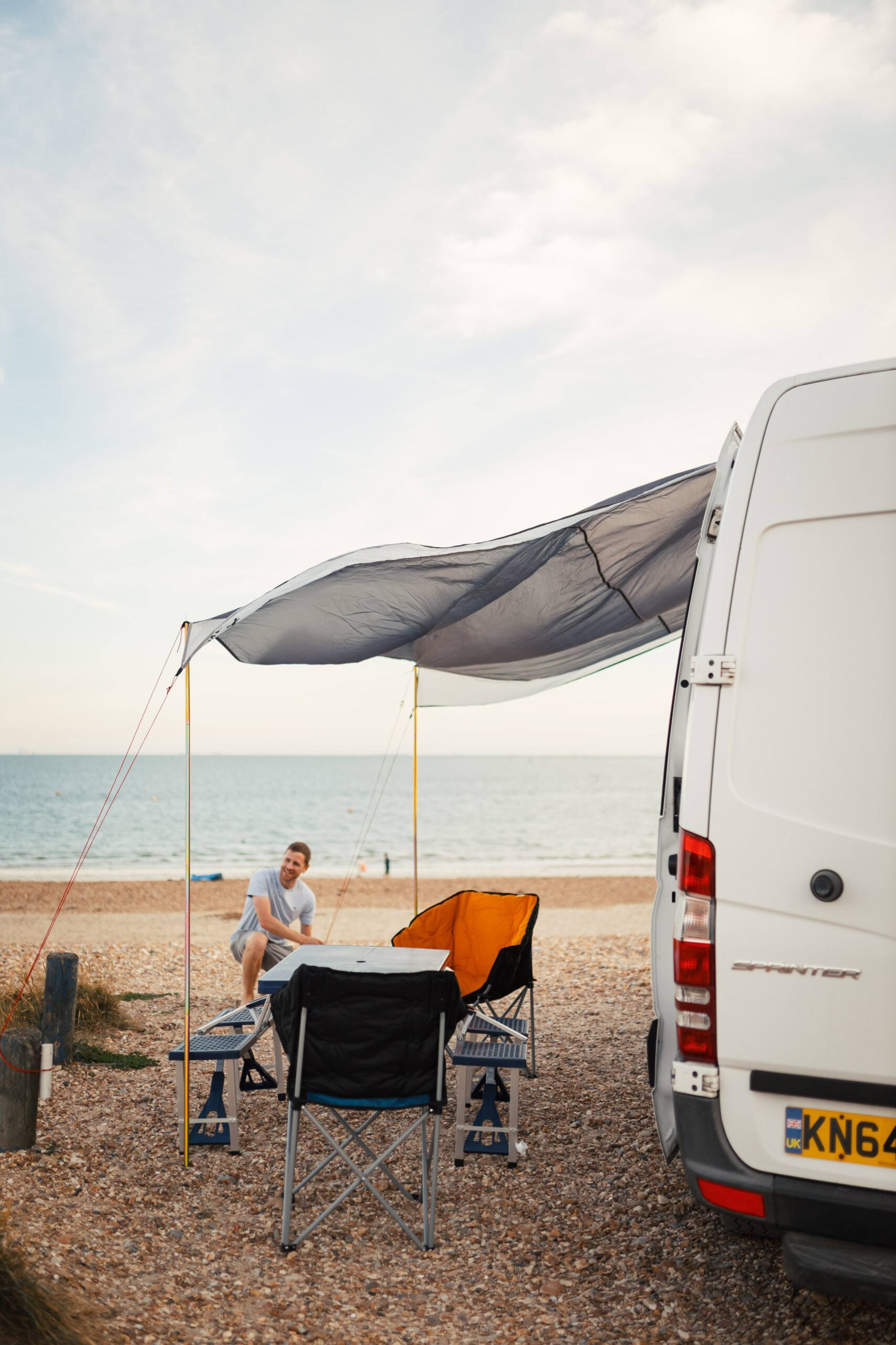 To the right is a campervan parked on a beach with it's awning set up. Under the awning is a picnic set up.