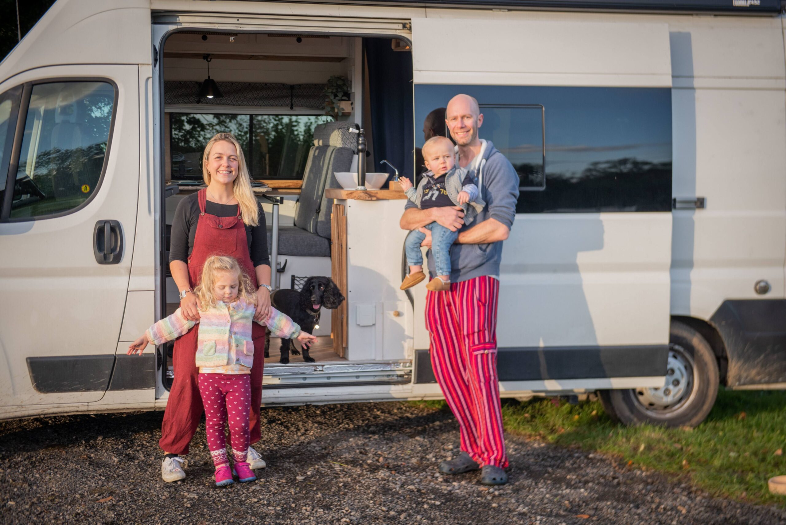 A young family of four stood smiling outside their campervan with the side door open